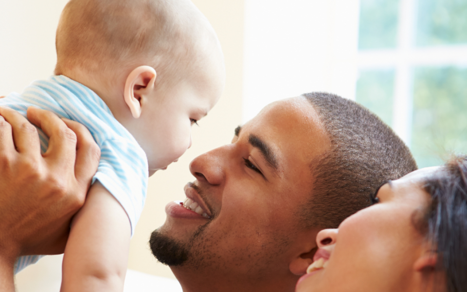 A man looking up at a baby in his hands, facing him, with a woman beside the man