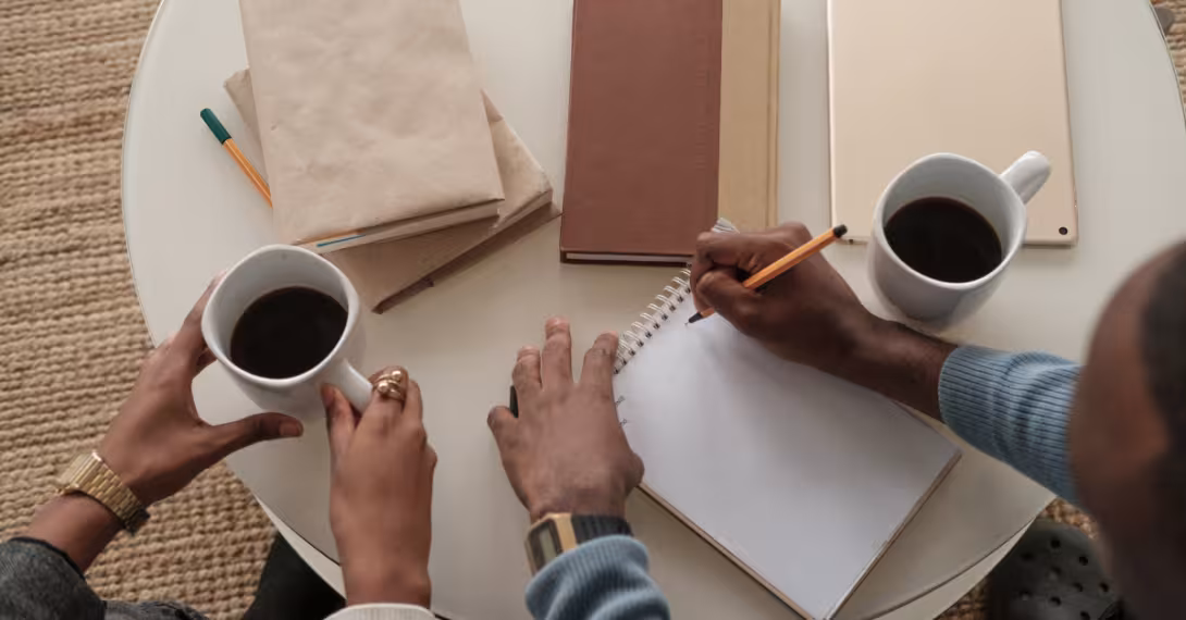 Hands over a table, one holding a cup of coffee and the other holding a pencil over a spiral notebook