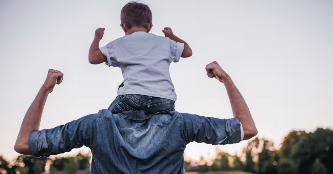 An adult, back turned, a child seated on their shoulders, both with arms lifted in the air