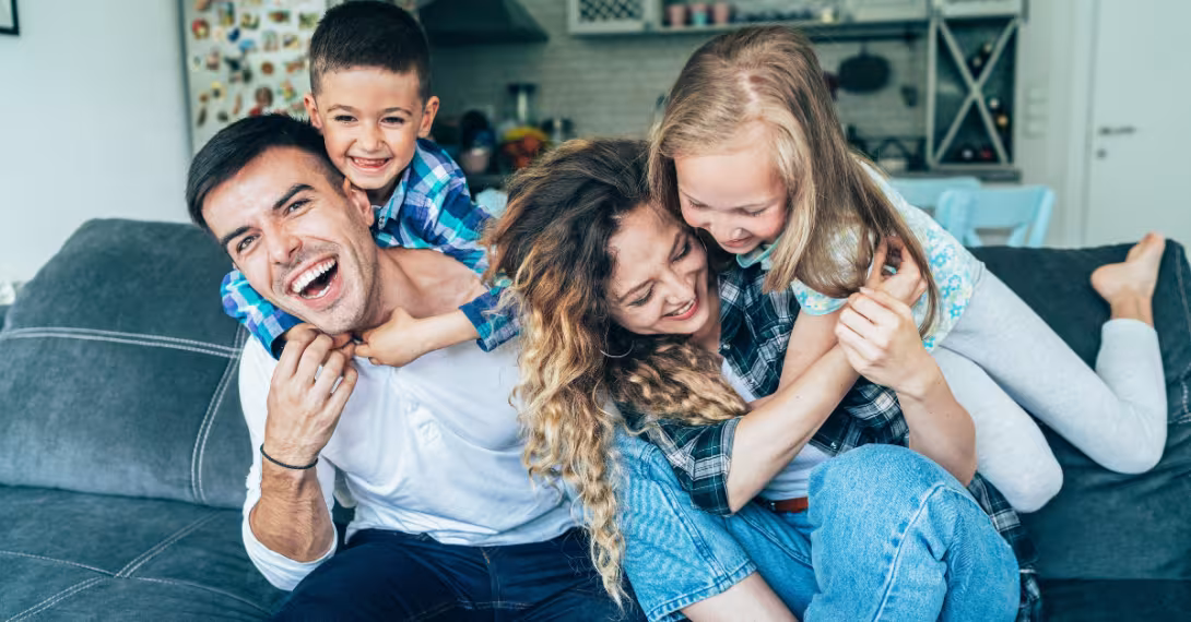 A family of four laughing on a couch as the children climb the backs of the adults